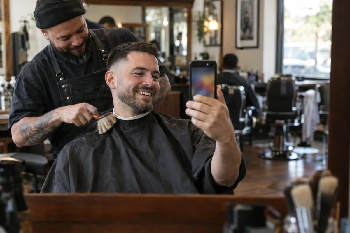A barbershop client records a video of his fresh haircut in the mirror while his barber finishes up