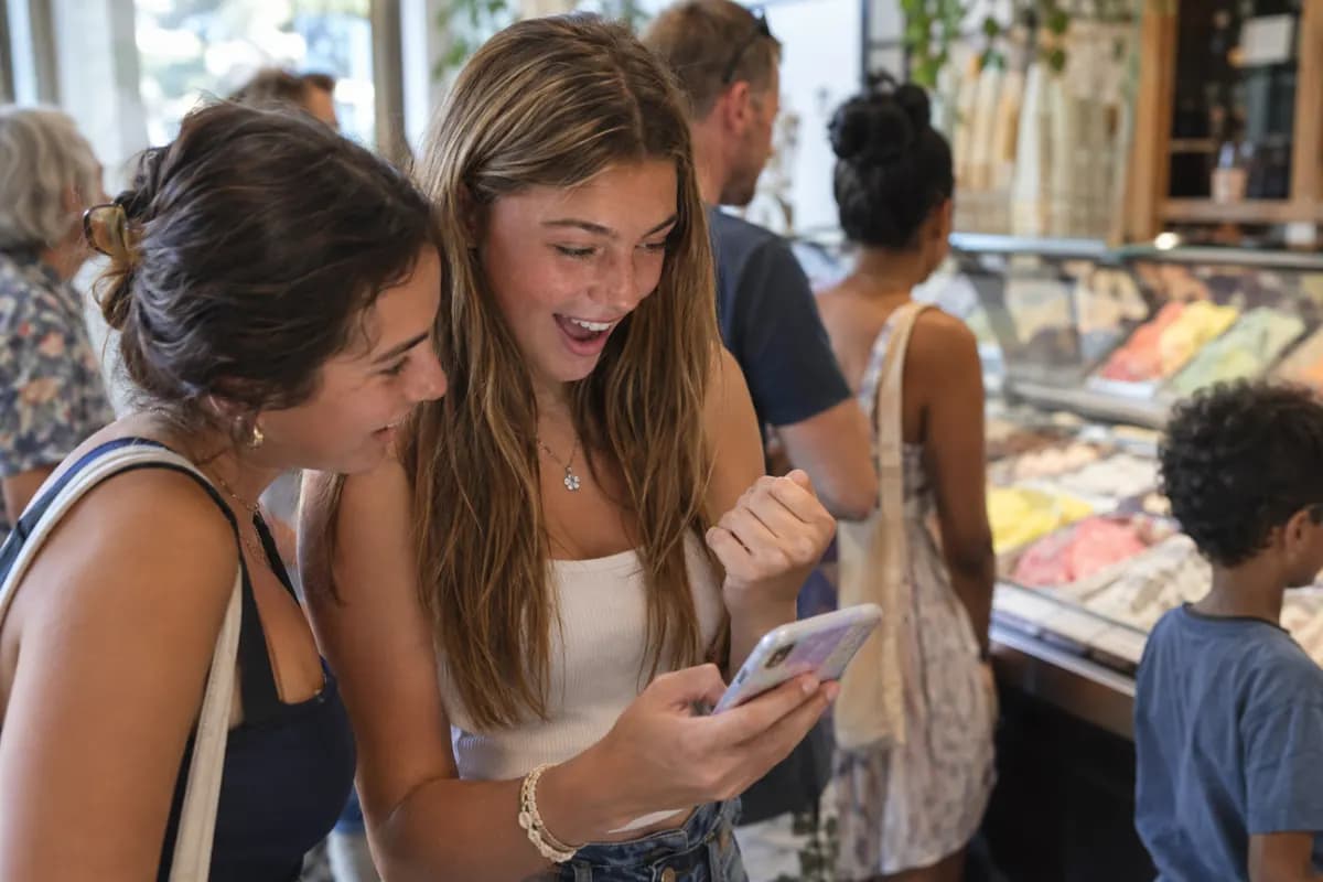 A teenager in an ice-cream shop queue reacts excitedly to her phone as a friend leans in