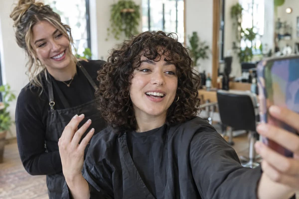 In a hair salon, a customer records a selfie video about her new haircut while her stylist smiles behind her
